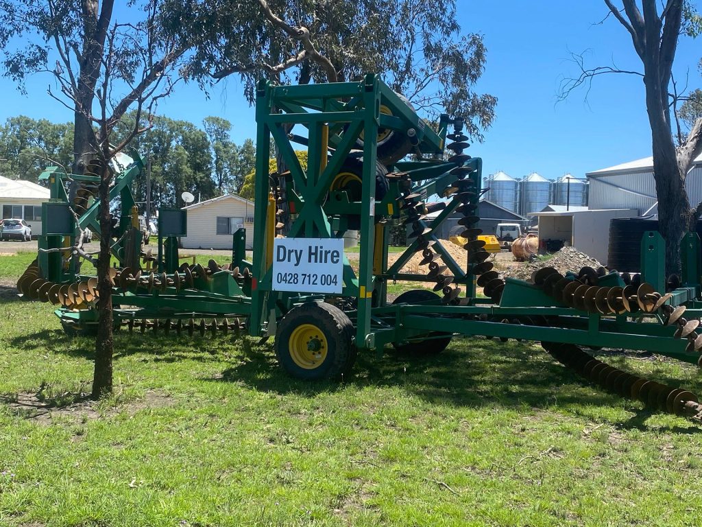 The image shows a large piece of green agricultural machinery parked on grass. There is a sign on the machinery that reads "Dry Hire" followed by a phone number. Trees and buildings are visible in the background.