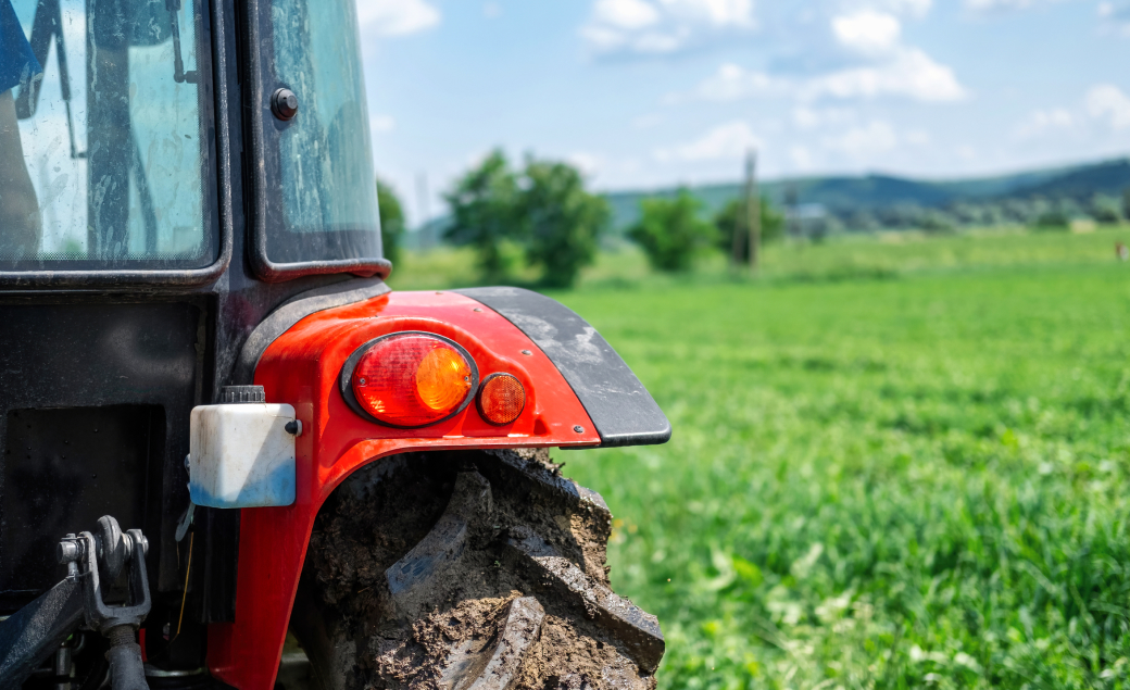 A close-up of the rear part of a red tractor in a green field, with a focus on the tire and rear light. The background shows a blurred landscape with trees and hills under a blue sky with clouds.
